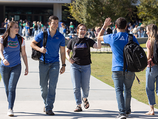 A group of five college students walk on a sunny campus path, smiling and chatting.