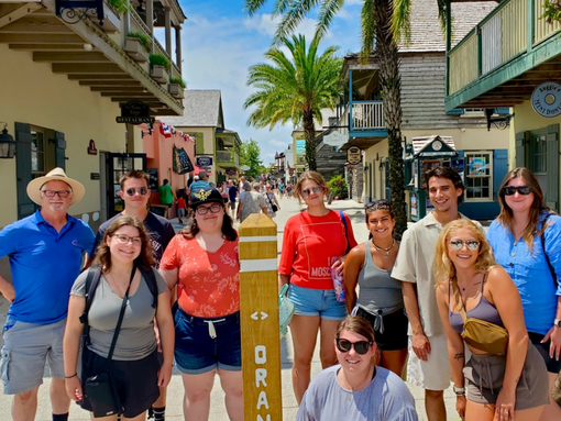A group of THEM students pose smiling on a sunny street lined with palm trees and shops